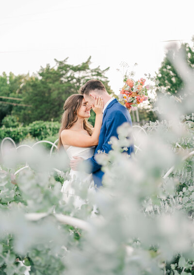 Bride and groom smiling at eachother in greenery with bride touching groom face