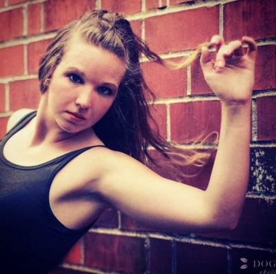 Woman standing against a brick wall with her left arm lifted and holding a piece of hair