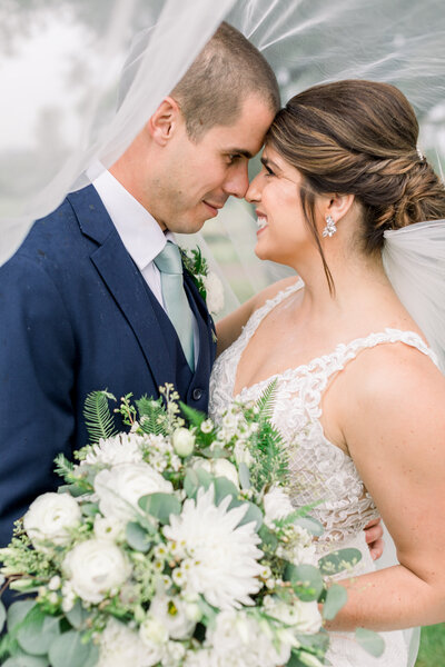 Bride and groom nose to nose smiling at each other with a bouquet under a veil