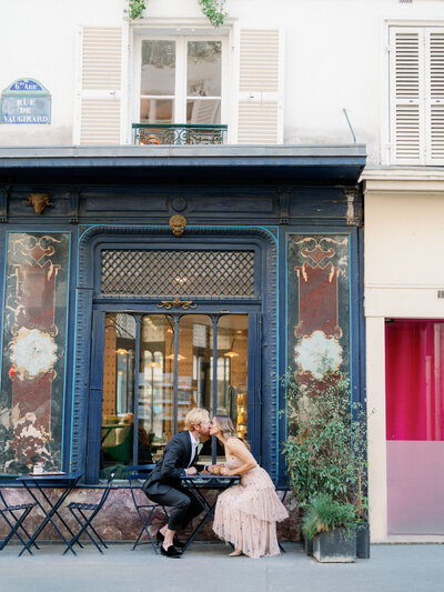 a man and woman nicely dressed kissing over a table at a corner cafe in Paris