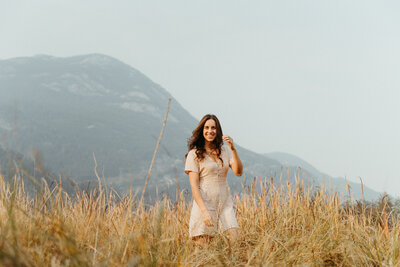Woman with brown curled hair wearing a beige dress in a field with a mountain in the background
