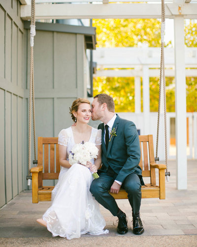 Couple sits on a swing as Groom kisses Bride on the cheek