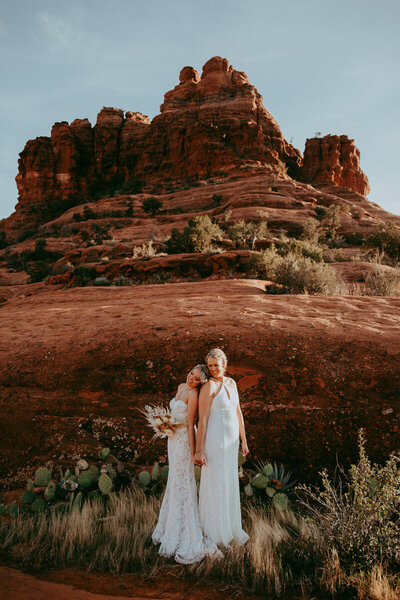 two women in white standing holding hands in front of bell rock in sedona arizona