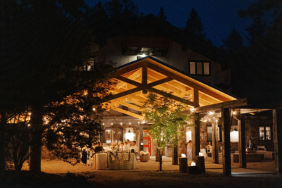 A rustic outdoor venue with a wooden pavilion adjacent to a white house with black trim. The pavilion is surrounded by trees and features a gravel pathway leading to the entrance at night.