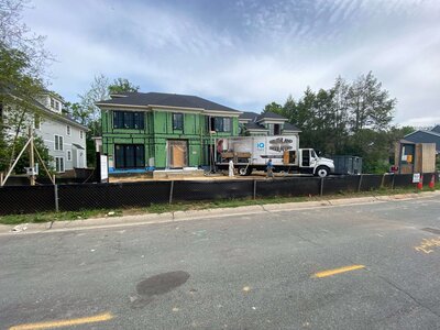 House under construction with exposed green insulation, construction materials, and a work truck parked in front, surrounded by a black fence.
