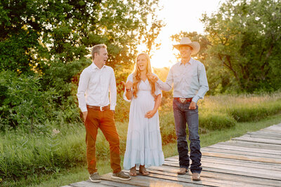 Three people stand on a wooden path in a sunlit park. A man and woman in casual outfits smile while a man in a cowboy hat joins them. Captured by Amber Bauer Photography, this Brainerd Lakes photography session features green trees with the sun setting in the background.