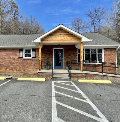 The office exterior is a dark red brick, ranch-style building. The porch reads "410" above a blue entryway door, framed by two potted plants. The door is accessible by two stairs leading up to the front door, as well as a ramp alongside the building, leading to the same door.