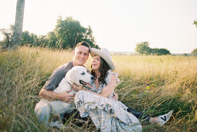 Couple sitting in a grassy field with their dog in their laps