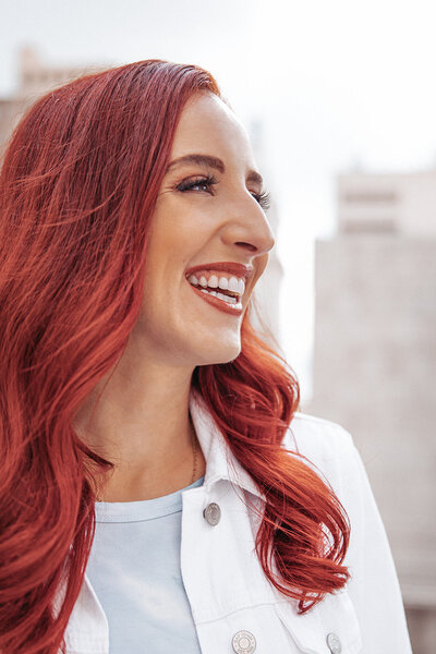 woman with red hair wearing a white jacket looks off to the right side while smiling