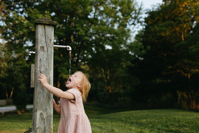 A child in a pink dress drinks water from an outdoor faucet on a wooden post, captured beautifully in the lens of a Toronto family photographer, set against the lush backdrop of a grassy park with trees.