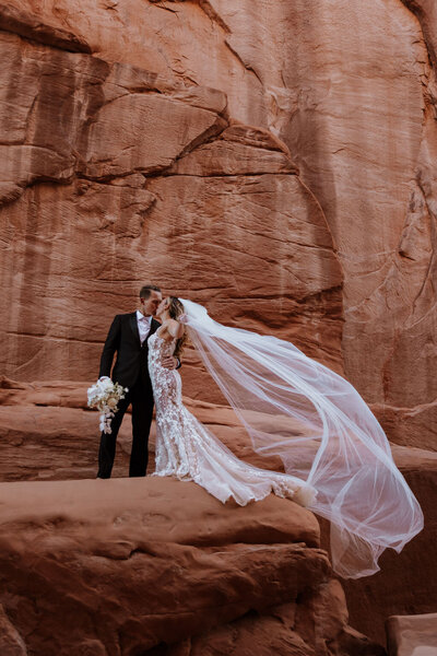 bride and groom kissing while standing on a large rock formation with a large walk wall behind them in the desert