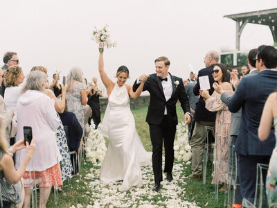 Joyful recessional at Wychmere Beach Club in Harwich Port, MA—bride raises bouquet as guests cheer and white petals line the aisle.