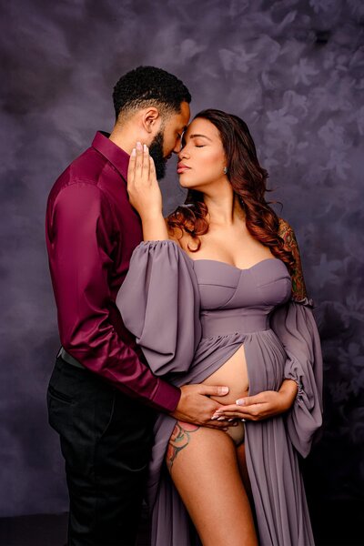 charlotte expecting couple is posed together during their maternity session in a waxhaw studio.  dad is wearing cranberry shirt and mom to be is wearing a purple dress