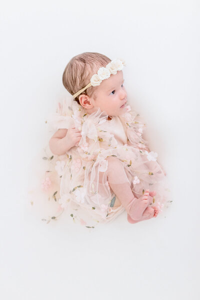 A baby dressed in a floral-patterned, frilly dress and wearing a flower headband sits against a white background, looking upward. The soft pastel colors create a delicate, ethereal atmosphere.