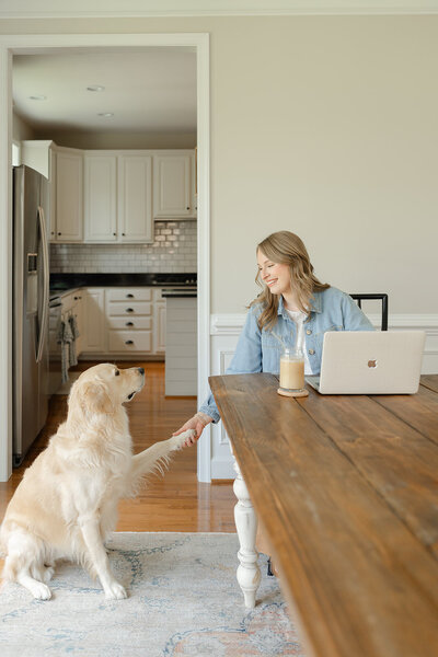 Woman leaning over a long wooden farm table to shake hands with a seated english-cream golden retriever