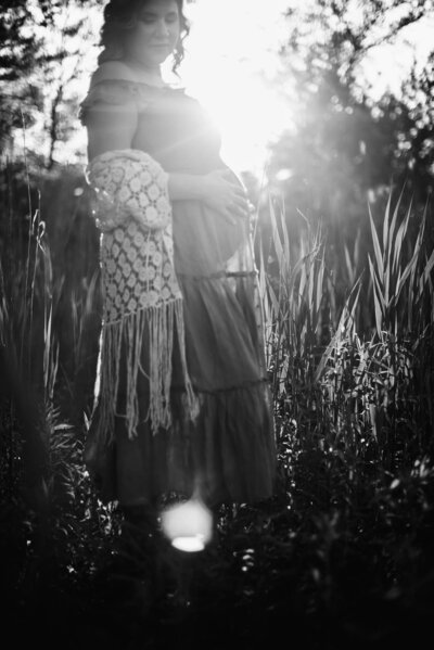 Maternity Photographer, Black and white image of woman standing in front of sunlight coming through the trees