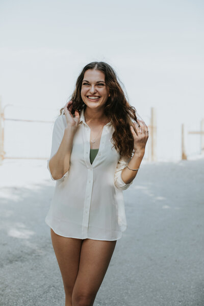 Woman dressed in a cream button down shirt laughing beside the beach.
