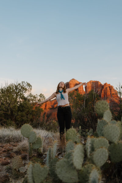 Brisa raises hands by prickly pear cactus with Sedona rocks behind.