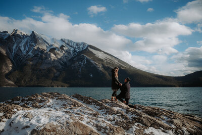 The photographer is crouched on a grassy hill with a lake behind her. She's holding a camera and looking to the right.