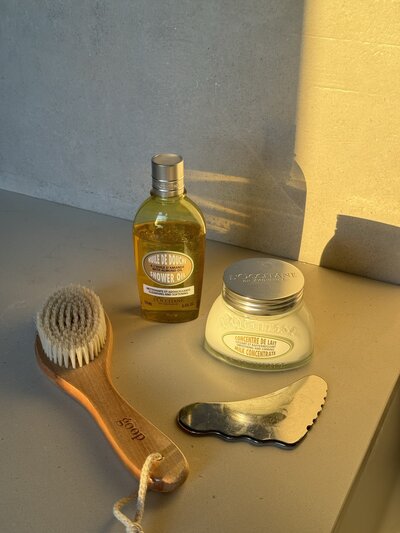 Natural light illuminates a shower oil, body cream, exfoliating brush, and metal tool on a beige surface, casting soft shadows, creating a warm, serene atmosphere.