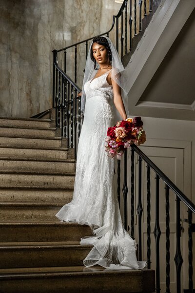 Bride in a white wedding dress with a veil, holding a colorful bouquet on a staircase at The Cadre in Memphis.