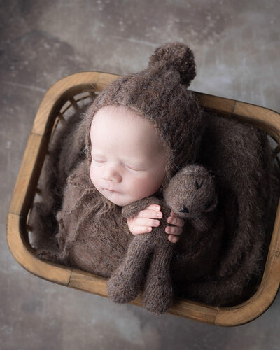 Sleeping newborn boy in fuzzy brown  wrap and bonnet, with brown background, holds small brown stuffed bear in both hands