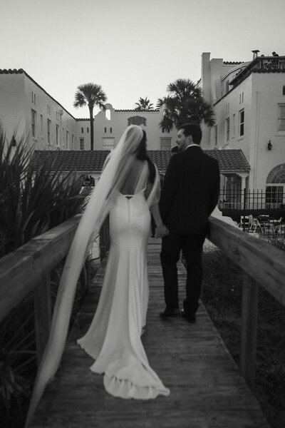 A black-and-white photo of a bride and groom walking hand-in-hand along a wooden boardwalk at Casa Marina Hotel in Jacksonville Beach, Florida. The bride’s sleek gown and flowing veil trail behind her, creating an elegant silhouette as the couple shares a quiet, romantic moment. Surrounded by Mediterranean-style architecture and palm trees, this timeless image was captured by Heartful Films, a Jacksonville wedding photographer and videographer known for emotional, cinematic storytelling.
