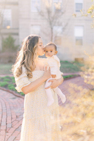 A young mother is holding her baby girl and giving her a kiss while standing outside