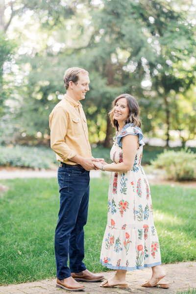 A couple holds hands and smiles, surrounded by lush greenery and dappled sunlight, creating a warm and romantic setting