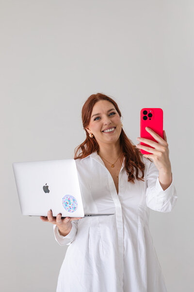 Mimi from Wed & Wilder wearing a white dress, holding a MacBook and red phone, with a smile.