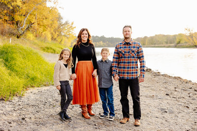 A family of four stands by a riverside in autumn, captured beautifully by a Brainerd Lakes Area Photographer. The woman wears a black top and orange skirt, the man sports a checkered shirt, while the children wear casual outfits. Fall foliage frames this serene moment alongside the calm river.