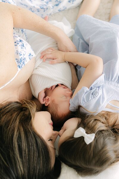 Mother in white and blue floral spaghetti strap dress holds her newborn between her and her toddler daughter as they snuggle noses, Indianapolis Photographer