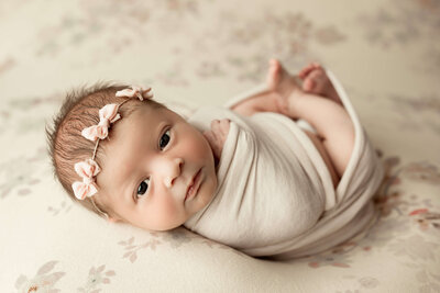 A serene newborn, captured during a Harrisburg newborn sessions, wrapped snugly in a soft blanket with a delicate floral headband, lying peacefully and gazing with innocent eyes.