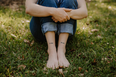 Brandi of the Intuitive Momma sits on the grass in jeans, showing off her "Be good, Do good" tattoos on her shins.