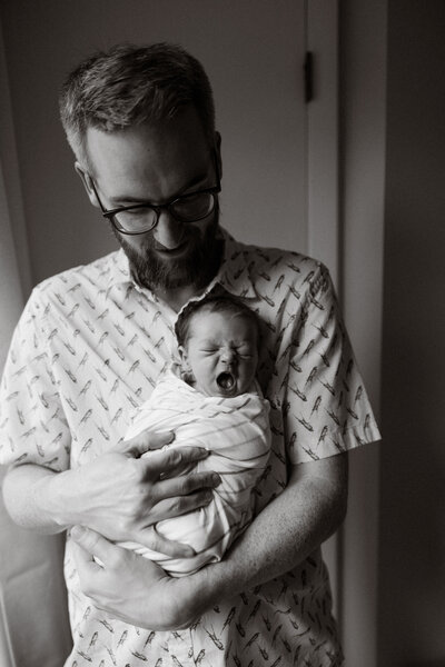 Adorable black and white newborn photography session of dad holding his baby girl, facing outward, yawning and swaddled, dad is looking down smiling