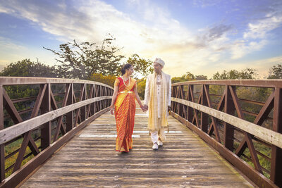 Indian wedding couple hold hands wearing traditional clothing and walk across bridge with dramatic sky in the background. Photo creatively captured by wedding photographer sacramento, philippe studio pro.