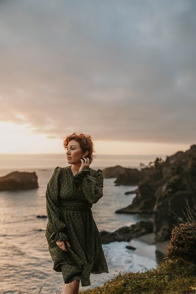 Woman with short red hair in a green dress stands on a cliffside at sunset overlooking the ocean, captured in the timeless style of an Ouray Colorado elopement photographer.