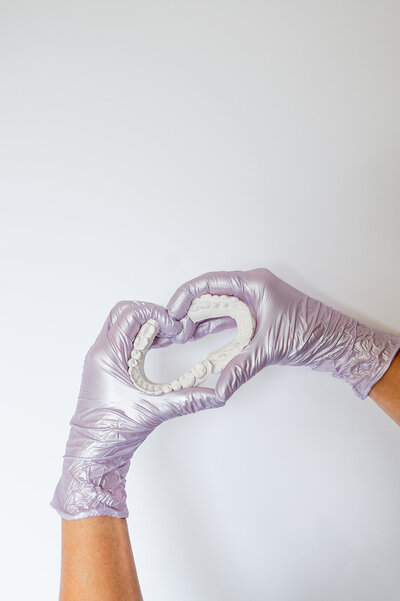 Two hands wearing shiny pink gloves hold two denture molds and form a heart shape.