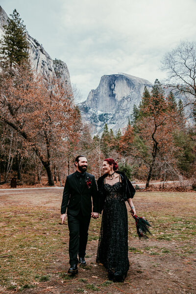Bride in a black dress and groom in a black suit walk hand in hand through Yosemite Valley with Half Dome in the background.