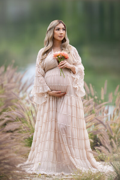 Pregnant mom standing in front of a lake in a long elegant sheer maternity dress holding her bump with one hand and some orange flowers in the other while staring off into the distance.