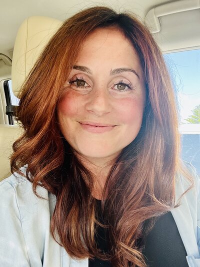 A woman with wavy auburn hair smiles softly while sitting in a car. Sunlight highlights her face, and she wears a black top with a light denim jacket.