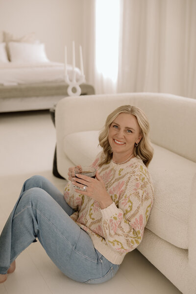 A female sitting on the ground, leaned against a couch drinking coffee smiling at the camera