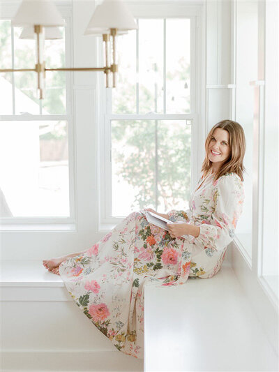 Meredith maag wears a floral dress smiling while seated on a bright window bench, softly lit by natural light – captured during a branding session in Kansas City.