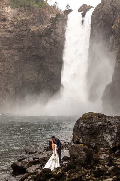 Bride and groom are having an adventure elopement standing beneath a massive waterfall and kissing