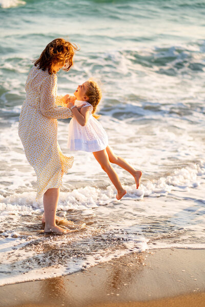 Woman in a yellow dress playing with a young girl at the edge of the ocean waves.