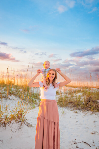 Mother holding her son on her shoulders with sand dunes and the purply colors of the sunset behind them.