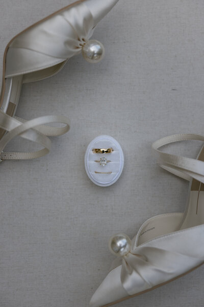 End-of-the-night wedding flat lay at Tree House Vero Beach, Florida, featuring a slice of white frosted cake on a woven charger with a gold fork, a bridal shoe with floral embellishments, “Groom” and “Bride” signs, wedding rings, tropical greenery, and a half-finished drink in warm candlelight.