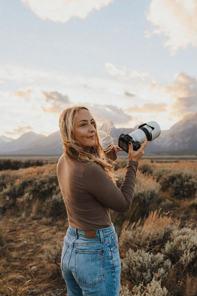 woman standing in an open field holding a camera with mountains in the background
