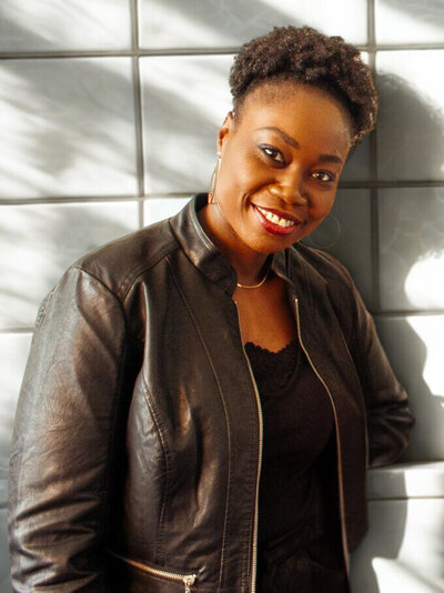 A professional portrait of a confident Black woman with shoulder-length twist braids and a warm smile. She is wearing a black turtleneck sweater and gray high-waisted pants. The background features a modern office environment with large windows showing an urban view.
