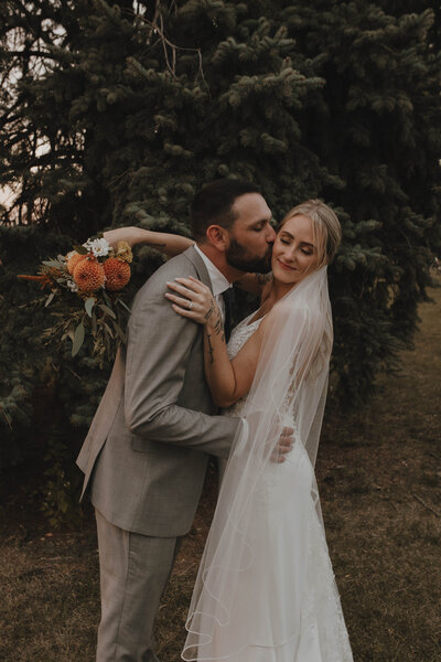 Groom kissing a brides cheek at an outdoor wedding in front of a tree